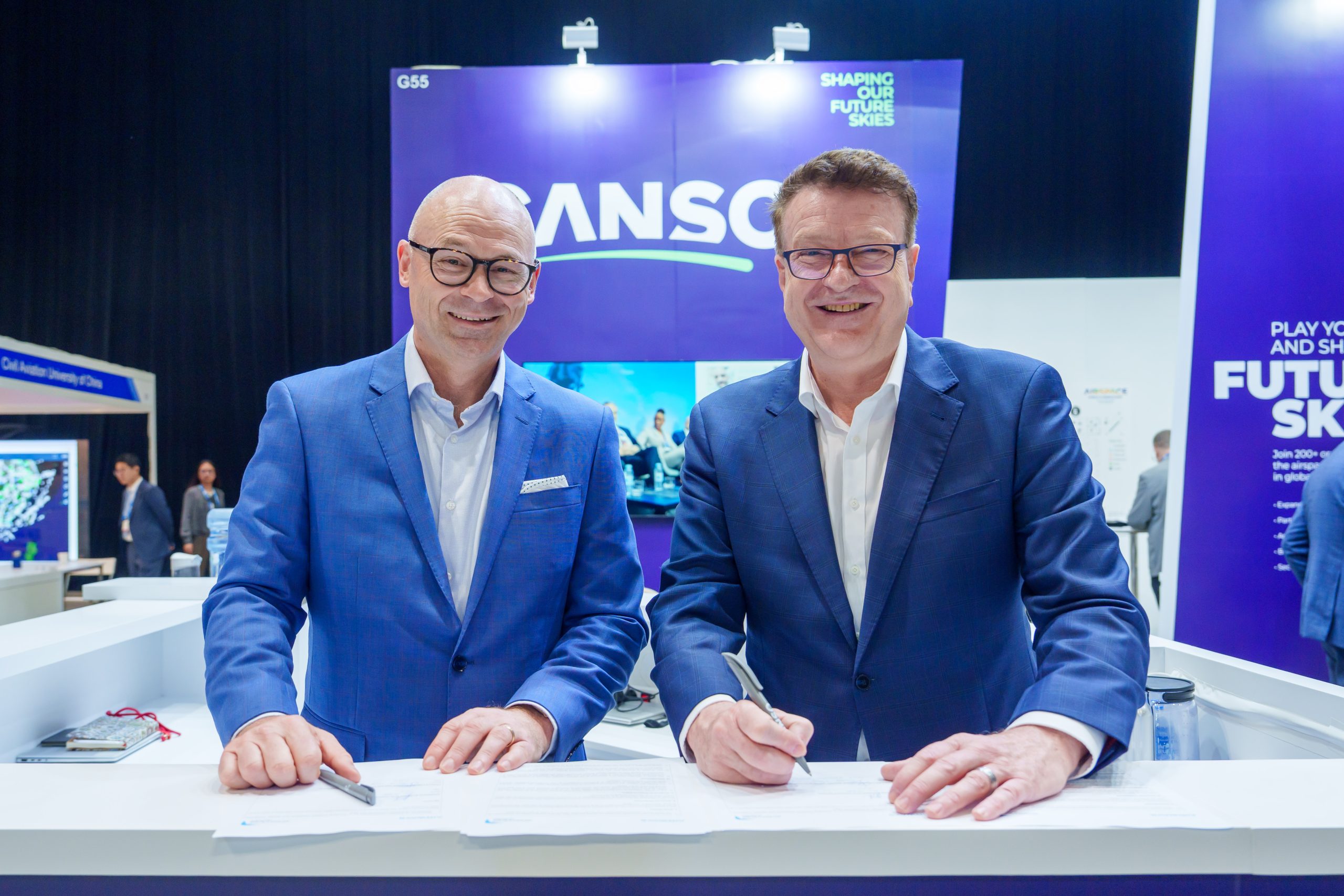 Two executives in blue suits signing an aviation safety and innovation agreement at an industry event.