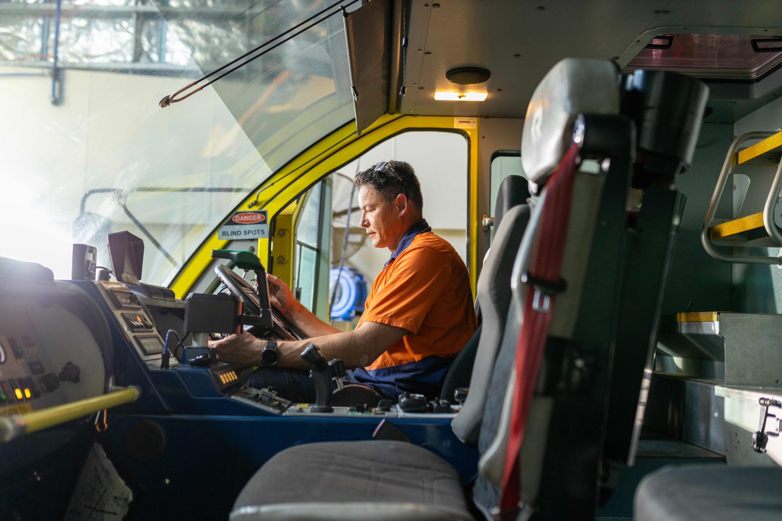 Person in an orange high-visibility shirt seated in a large vehicle cabin, operating the steering wheel and controls.