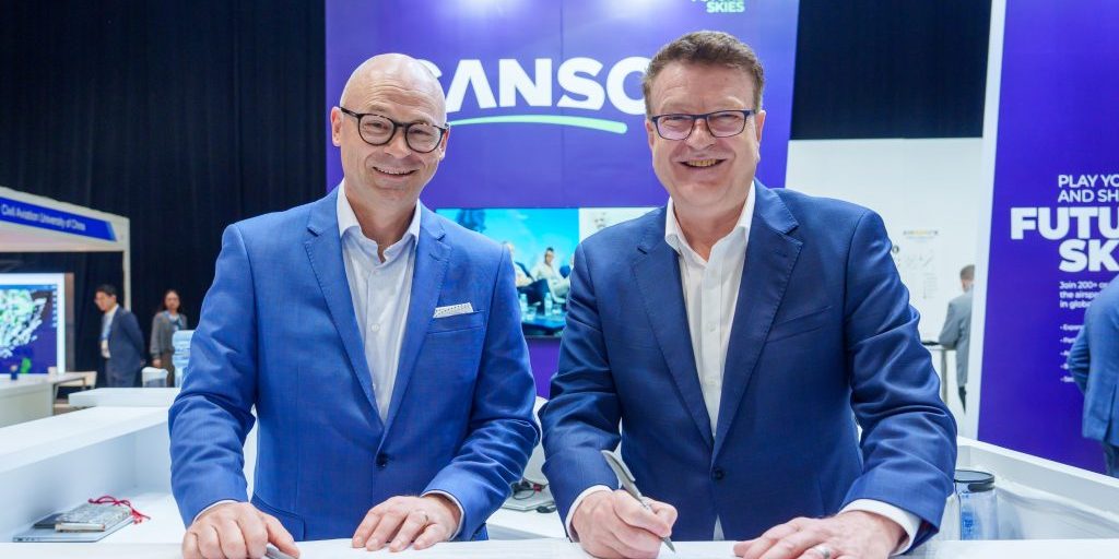 Two executives in blue suits signing an aviation safety and innovation agreement at an industry event.