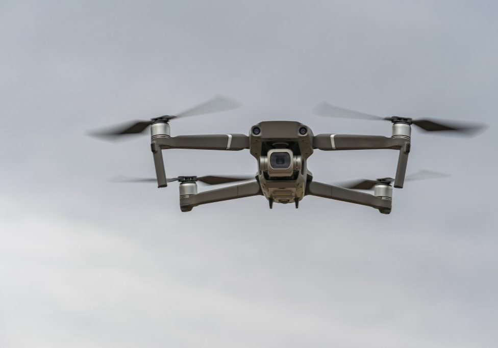 A small quadcopter is flying at a low altitude against a gray sky and the camera is aimed at the photographer.